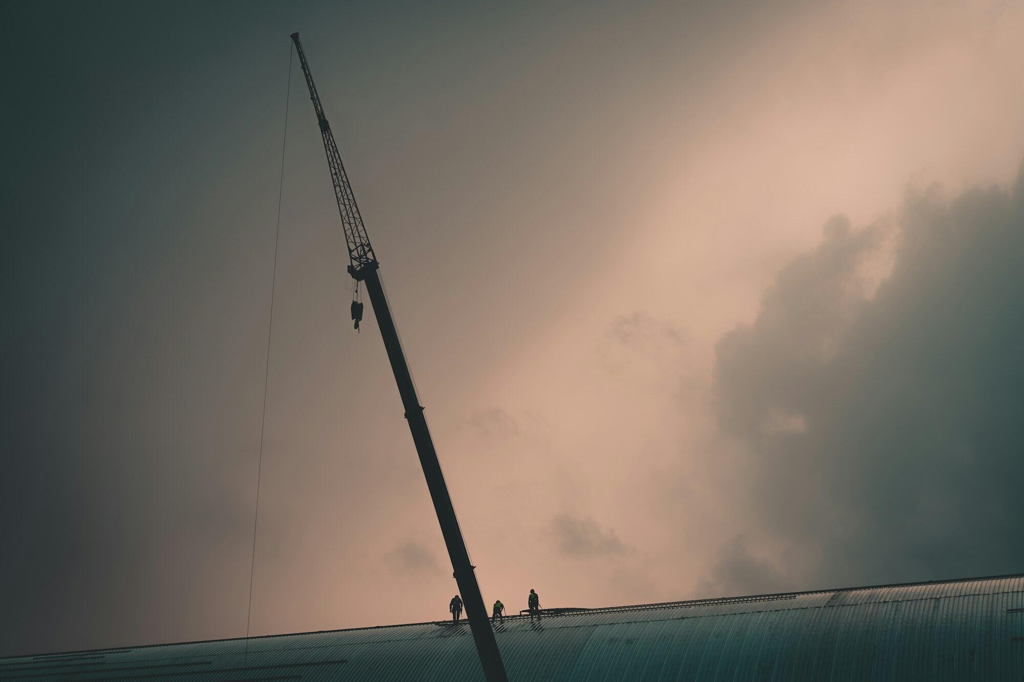 A crane boom and three workers on a roof as storm cloud clears.