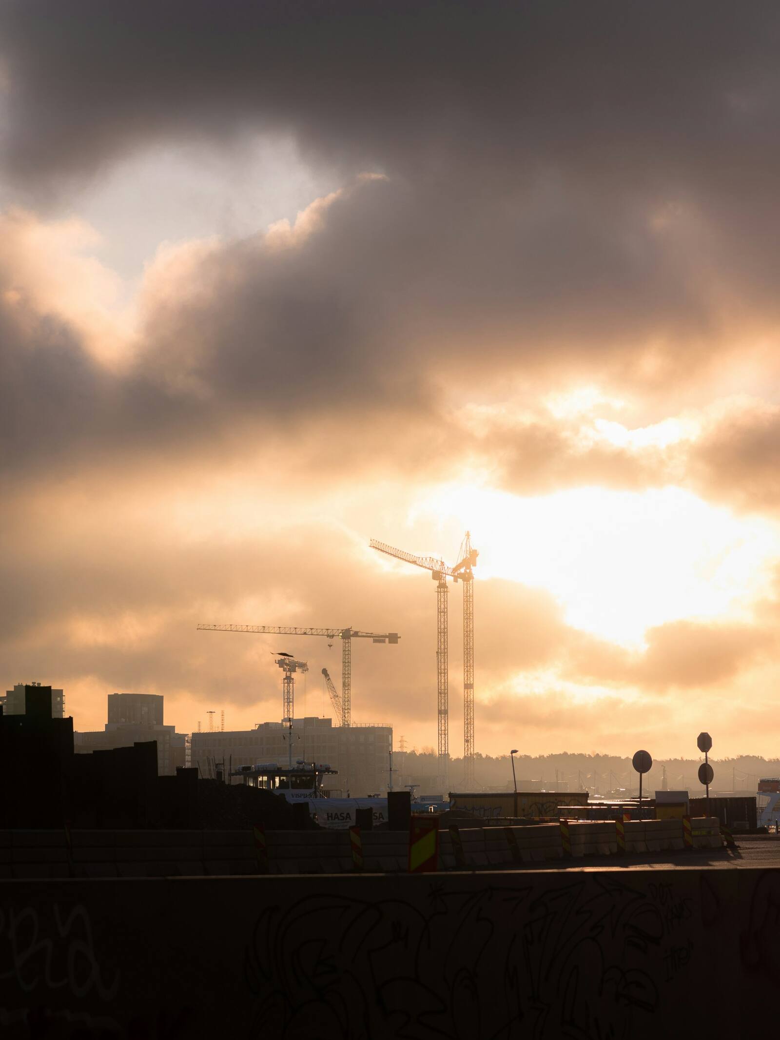 Tower cranes over a city site as the sun drops behind cloud.
