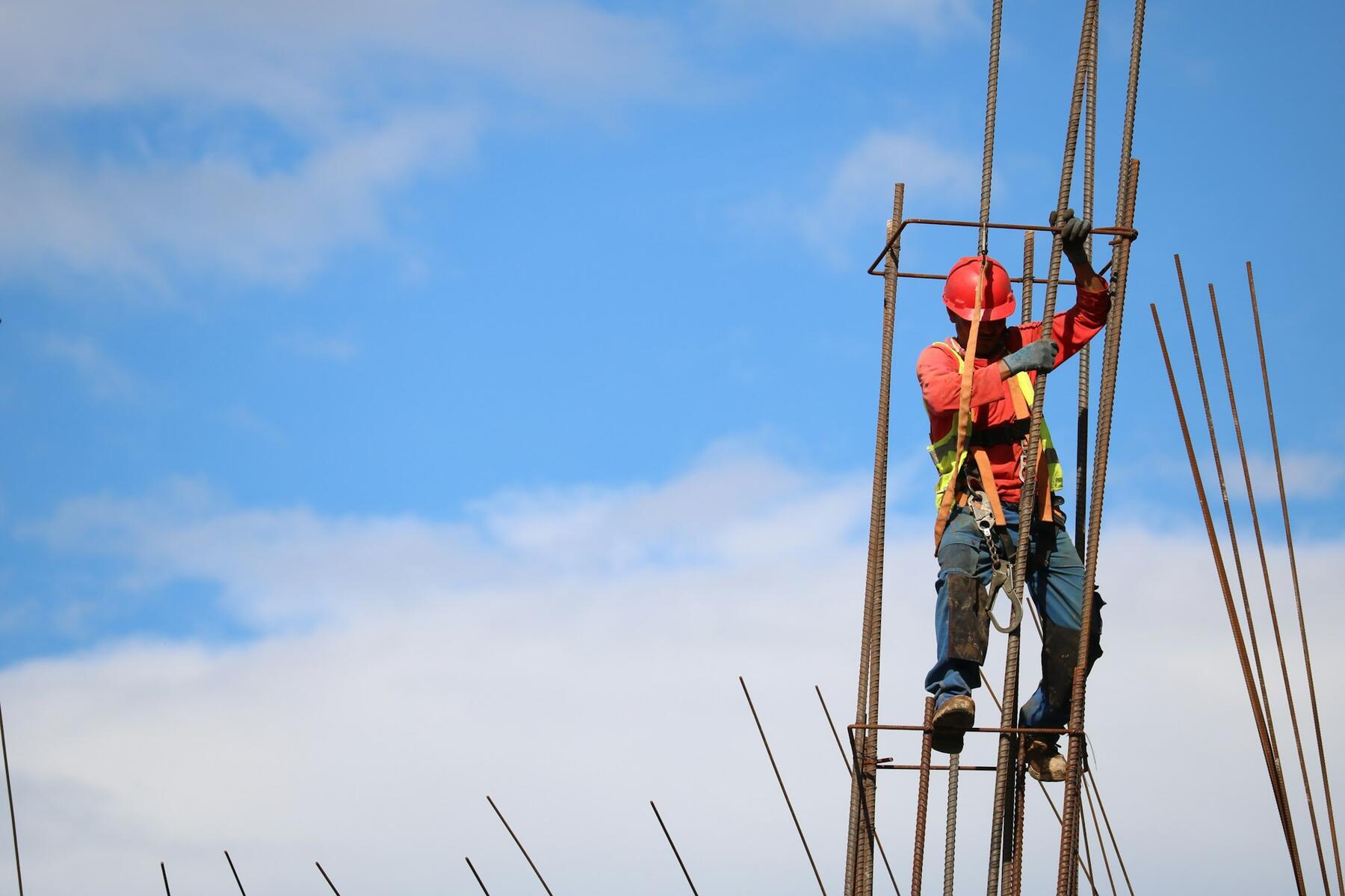 A worker in a red hard hat climbing a rebar cage against a blue sky.